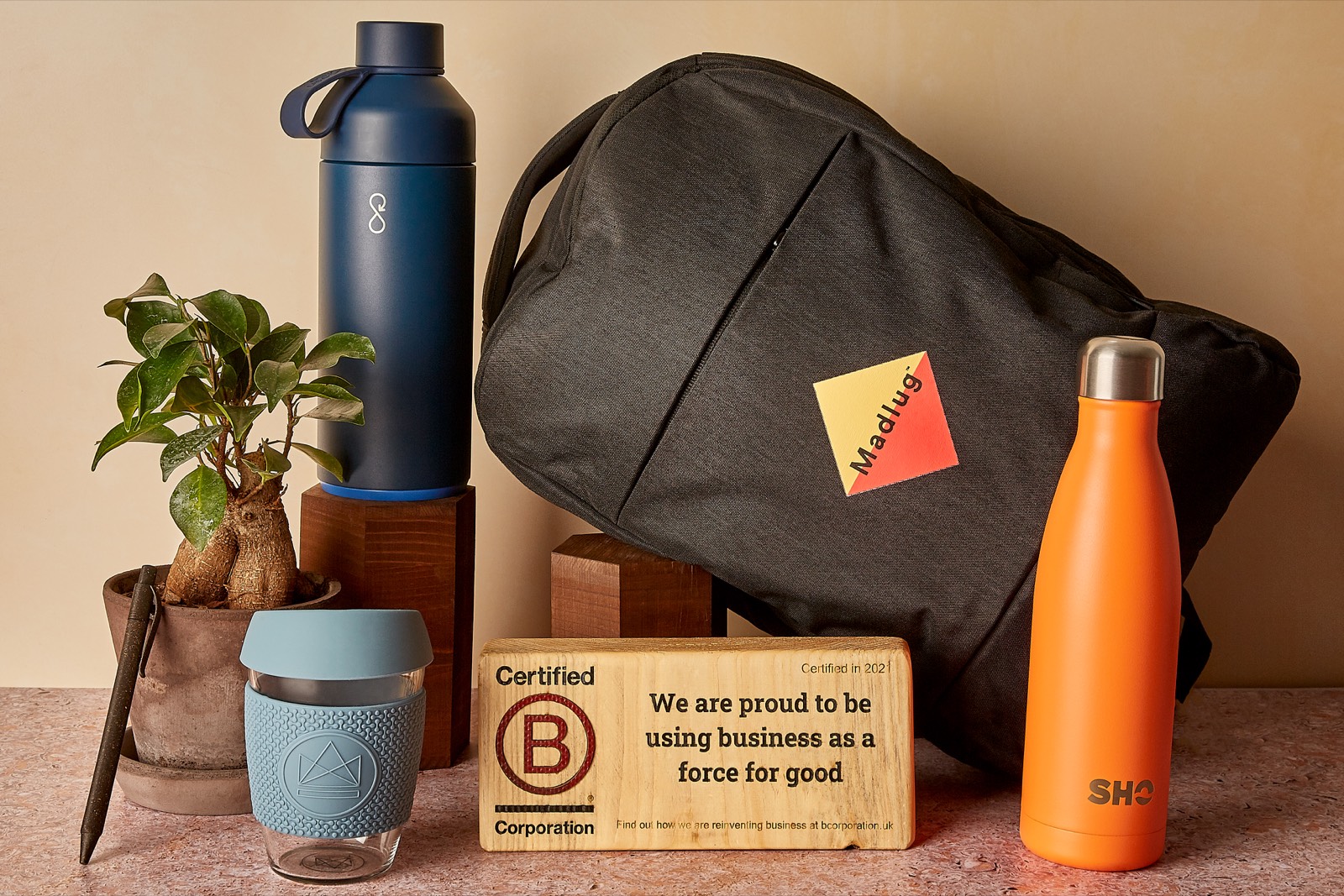 Branded merchandise on a shelf: a navy Ocean Bottle, a Madlug backpack, an orange SHO bottle and a reusable coffee cup, beside a wooden Certified B Corporation plaque